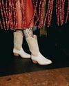 Person in white cowboy boots and tattooed legs under a red skirt stands next to a red beaded curtain on a wooden floor.