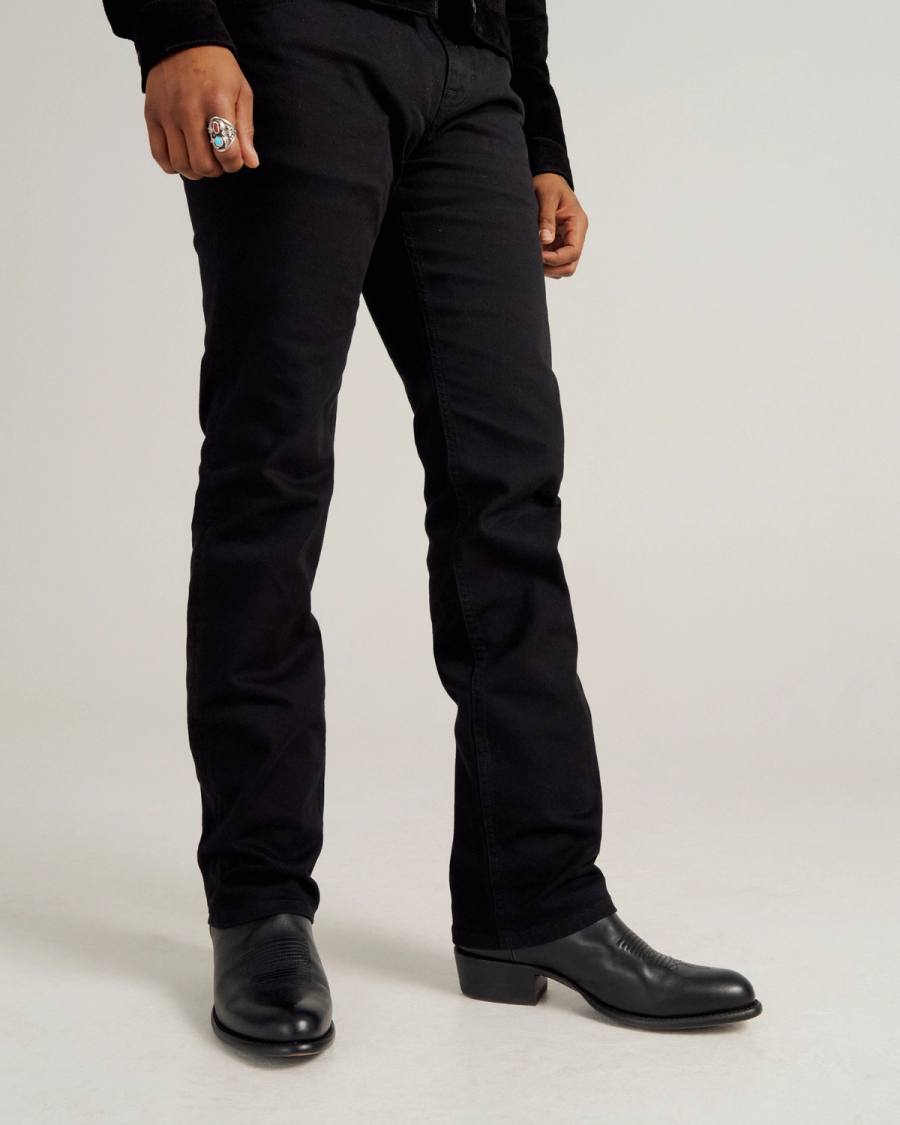 Person wearing black pants, black boots, and a ring, standing against a plain light background. Only the lower body is visible.