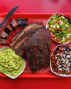 A red tray holds grilled ribs, sliced brisket, and burnt ends, accompanied by guacamole pasta, diced avocado salad, and a black bean and corn salad with crumbled cheese. Plastic cutlery is placed on the tray.