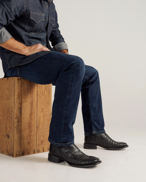 The William in Midnight Caiman. A person wearing a denim shirt, dark blue jeans, and black Midnight Caiman cowboy boots sits on a wooden box against a plain background.