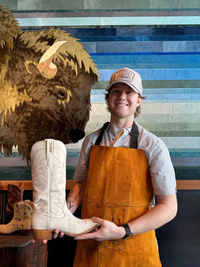 A person wearing an apron and a cap holds a white cowboy boot in front of a buffalo-themed wall art. Other boots are displayed on a wooden shelf to the left.