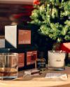 A glass of whiskey, three stacked Ranger Station boxes, a perfume bottle, a candle, and matches are arranged on a table near a decorated Christmas tree.