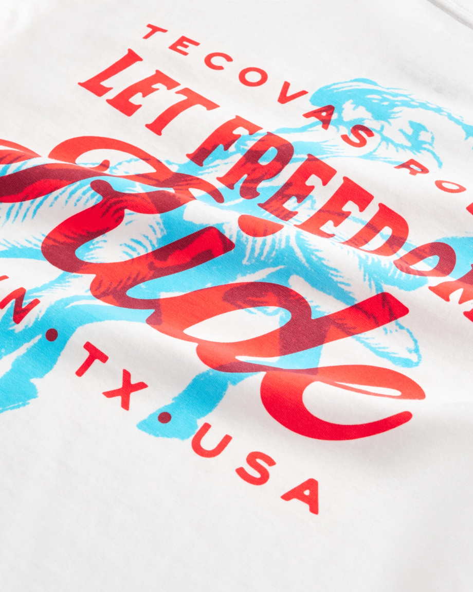 White t-shirt with red and blue graphic text reading “Tecovas presents Let Freedom Ride” and “ATX, TX, USA,” featuring a cowboy on a horse illustration.
