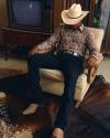 Person in a cowboy hat, snakeskin shirt, and boots sits in an armchair on an animal hide rug, with an old TV and cabinet in the background.