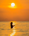 Silhouette of a person fishing in the sea during a vibrant orange sunset, with the sun low in the sky and birds flying in the background.