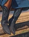Person wearing blue cowboy boots and a blue skirt sits on a wooden fence with fields in the background.