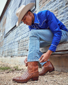 Man in cowboy attire adjusting his boot on an urban street.