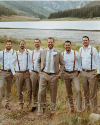 Six men pose outdoors by a lake and mountains; one wears a tan suit with a tie, while the others wear white shirts with suspenders and tan pants.