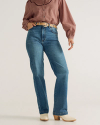 Person wearing a mauve long-sleeve blouse, blue high-waisted jeans, a beige belt, and white heeled boots, standing against a plain background.