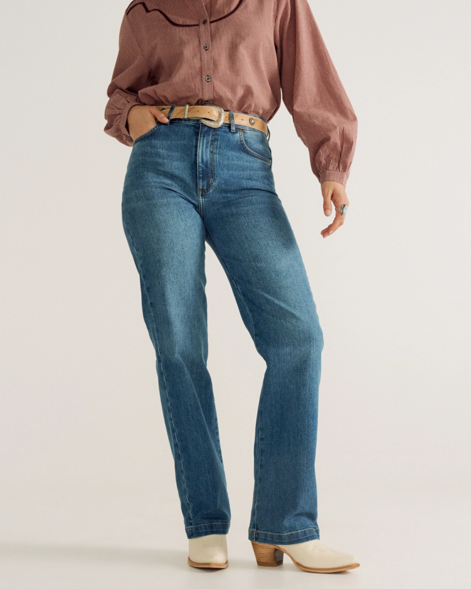 Person wearing a mauve long-sleeve blouse, blue high-waisted jeans, a beige belt, and white heeled boots, standing against a plain background.