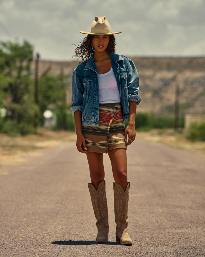 The Abby in Driftwood Suede. A woman stands on a rural road in The Abby Suede cowboy boots, paired with a denim jacket, white tank top, and patterned skirt. Trees and a mountain are visible in the background.