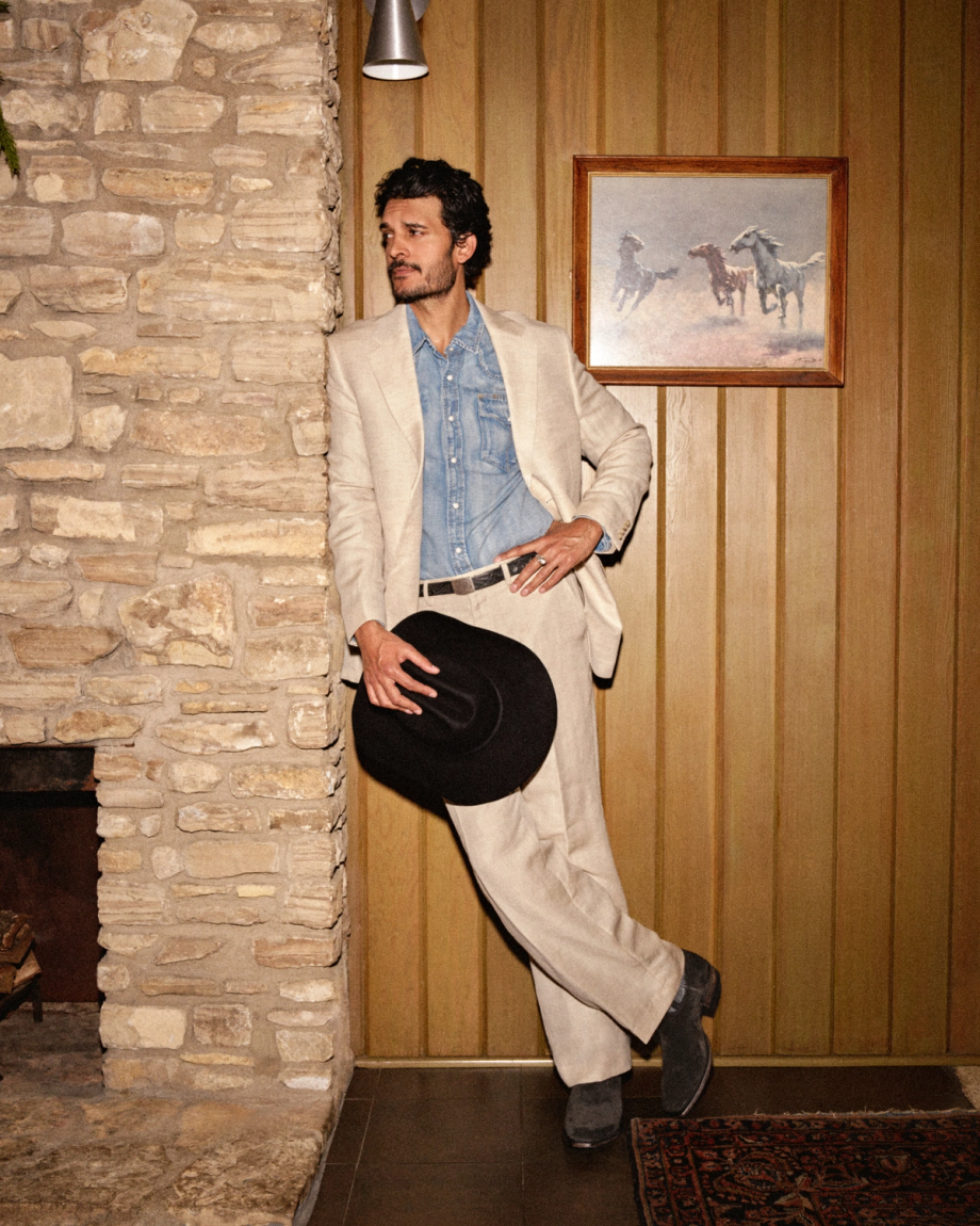 The Warren in Gunsmoke Suede. A man in a beige Warren Suede suit and denim shirt leans against a stone fireplace, holding a black cowboy hat.