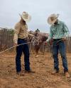 Two people in cowboy hats stand on dry ground, adjusting ropes. A saddled horse stands in the background against a wooden fence.