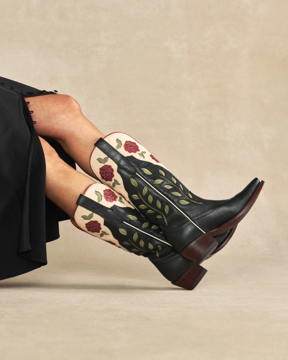 The Annie in Midnight Roses Cowhide. Person wearing black dress and Midnight Roses Cowhide cowboy boots with red rose and green leaf embroidered designs, seated on a neutral background.
