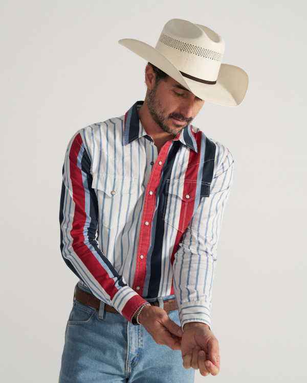 A man wearing a cowboy hat and a striped western shirt is looking down while adjusting his shirt cuff.
