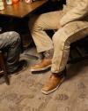 Two people sitting at a wooden table, wearing work clothes and boots. Floor has visible footprints and dirt.