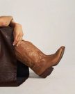 The Britt in Latte Goat. A person wearing brown cowboy boots sits with legs crossed, partially covered by a dark brown draped fabric, while a curious goat stands nearby.