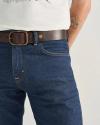 A person wearing blue jeans, a brown belt, and a white shirt, with a small sun tattoo visible on their inner forearm.