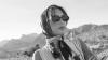 A woman wearing sunglasses and a headscarf stands in a rocky desert landscape with mountains in the background, looking away from the camera.