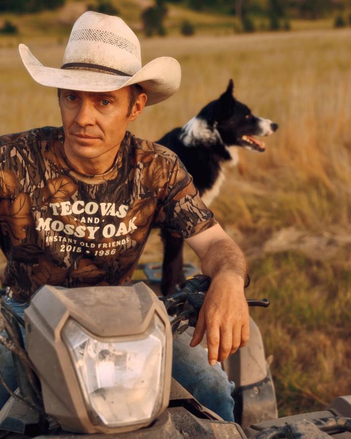 A man in a cowboy hat sits on an ATV in a field, wearing a camo shirt, with a black and white dog sitting behind him.