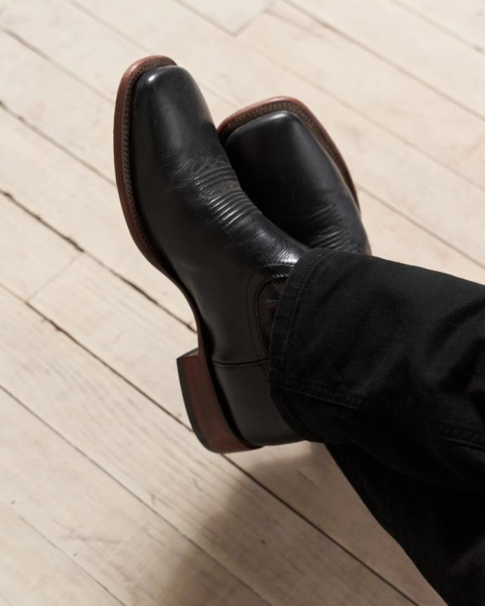 A person wearing black leather boots with brown soles is shown crossing their legs on a light-colored wooden floor.