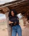 A person with curly hair in a black sweater and jeans leans against a wooden post on a rustic porch.