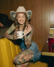 Woman with tattoos and cowboy hat sits on a yellow booth, holding a mug and smiling. Brown wood-paneled wall in background.