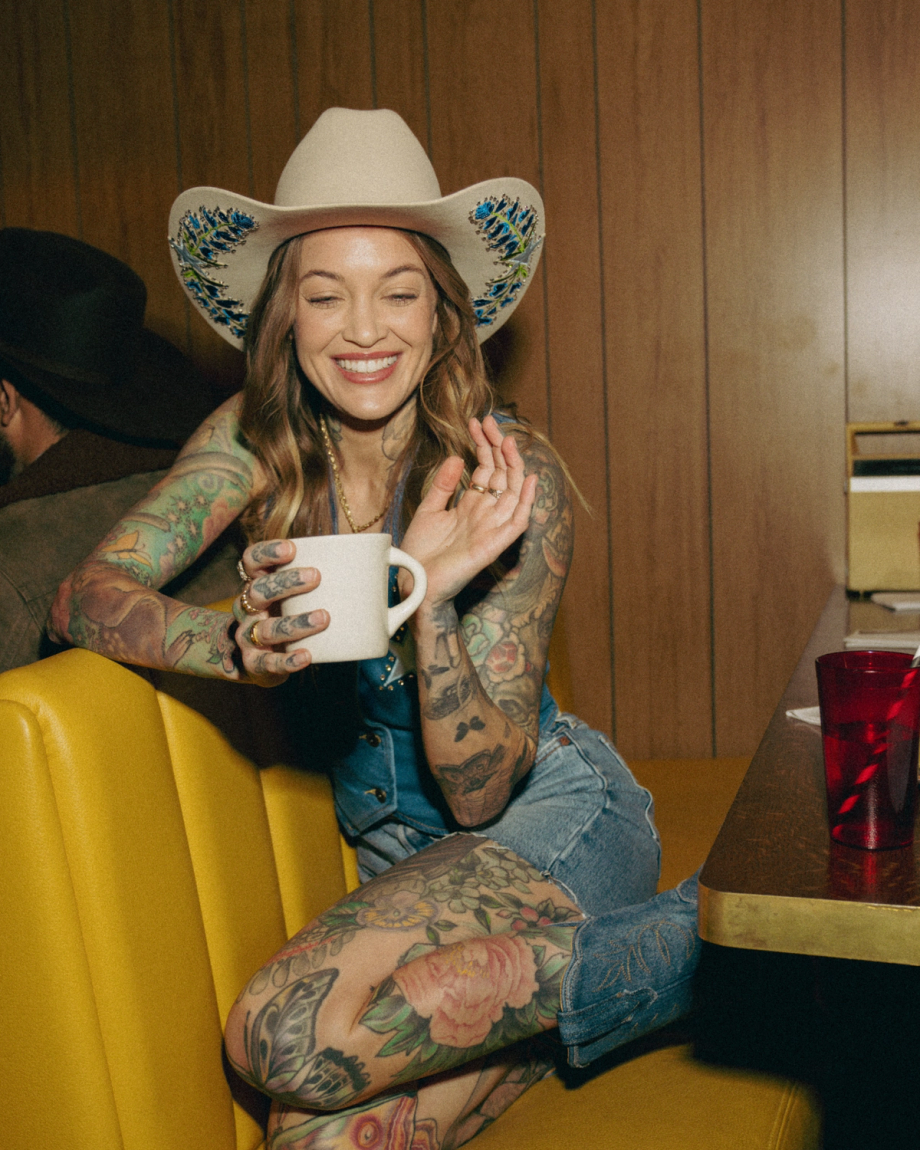Woman with tattoos and cowboy hat sits on a yellow booth, holding a mug and smiling. Brown wood-paneled wall in background.
