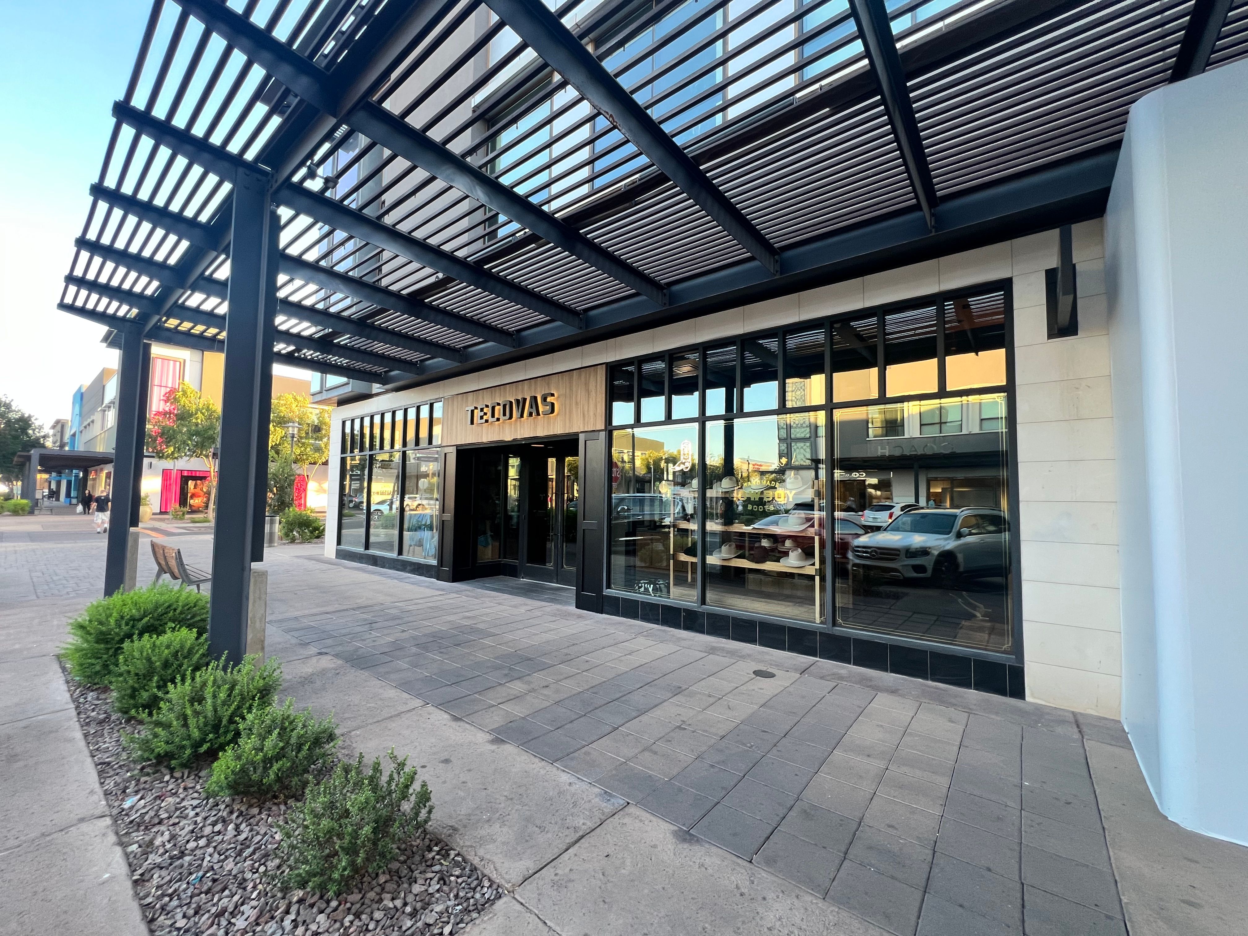 Exterior of a Tecovas store with large windows and a black metal awning, showing parked cars reflected in the glass and landscaped sidewalk in front.