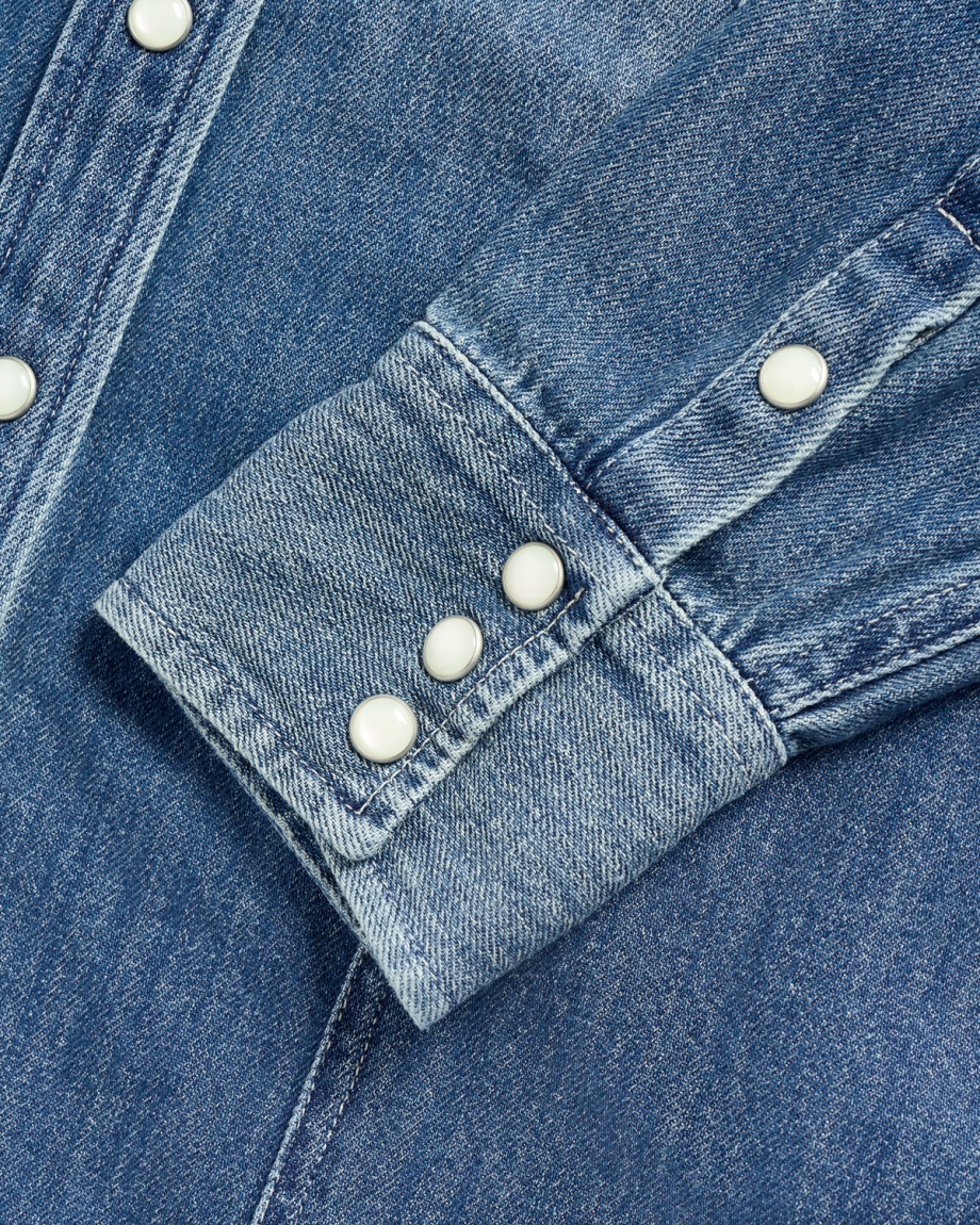 Blue denim shirt sleeve with three white snaps on the cuff and one on the placket.