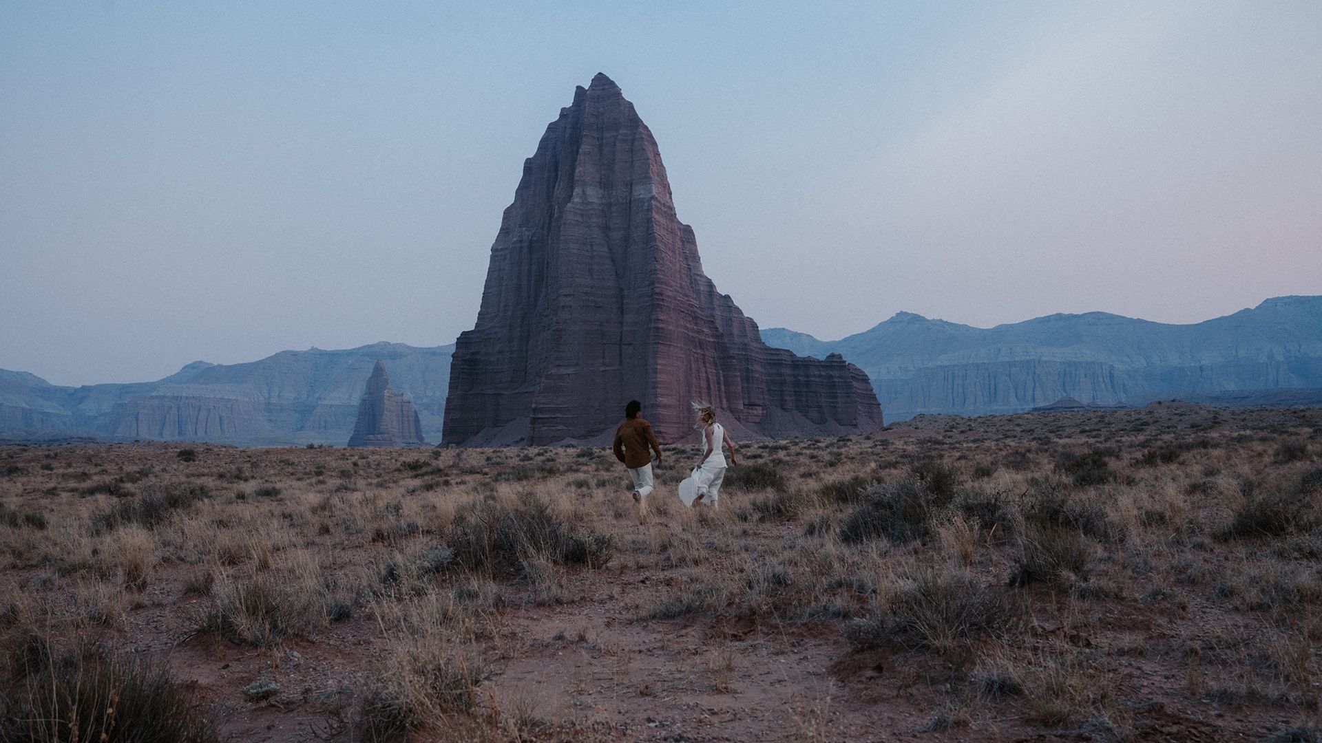 A couple walks through a desert field toward a large, jagged rock formation.