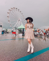 A woman in a dress and cowboy hat poses with a Ferris wheel and various booths in the background at a fair on a rainy day.