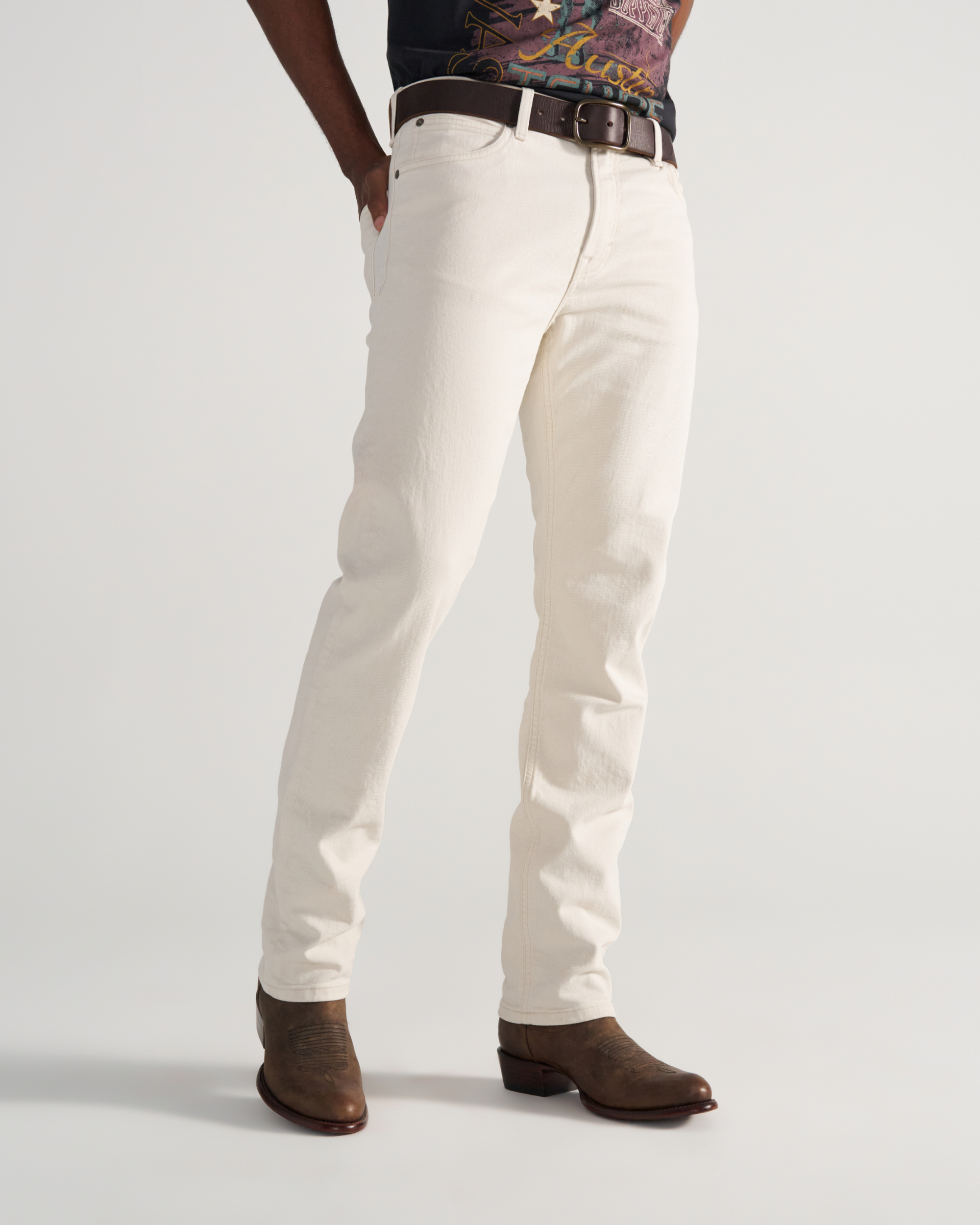 Person wearing off-white jeans, a brown belt, a graphic t-shirt, and brown cowboy boots, standing against a plain light background.