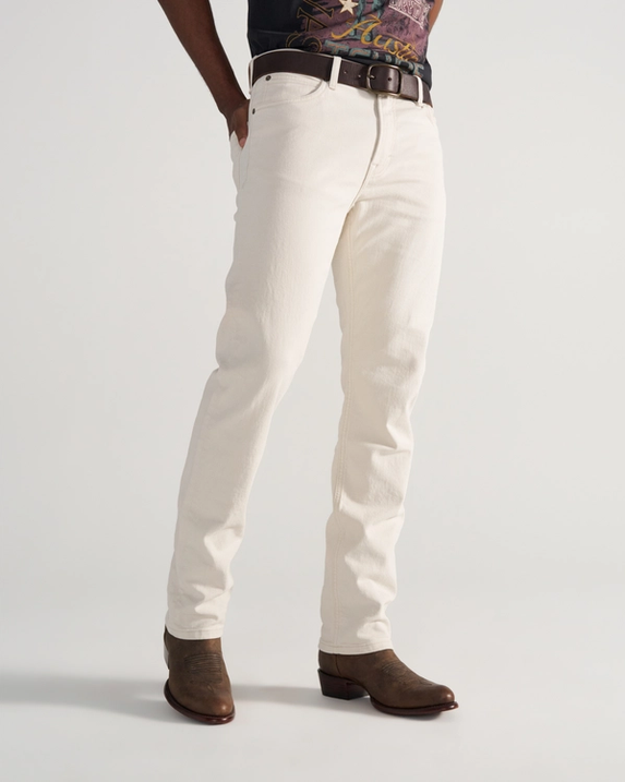 Person wearing off-white jeans, a brown belt, a graphic t-shirt, and brown cowboy boots, standing against a plain light background.