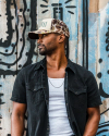 A man wearing a black shirt, white tank top, and camo hat with "MAS" on it looks to his left while standing against a painted wooden fence background.