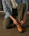 Person kneeling on one knee, tying the laces of a women's work tan boot, wearing olive green pants, a white shirt, and a light blue denim jacket.