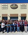 A group of people stands in front of a Murdoch's Ranch & Home Supply store, holding a large ribbon for a ribbon-cutting ceremony.