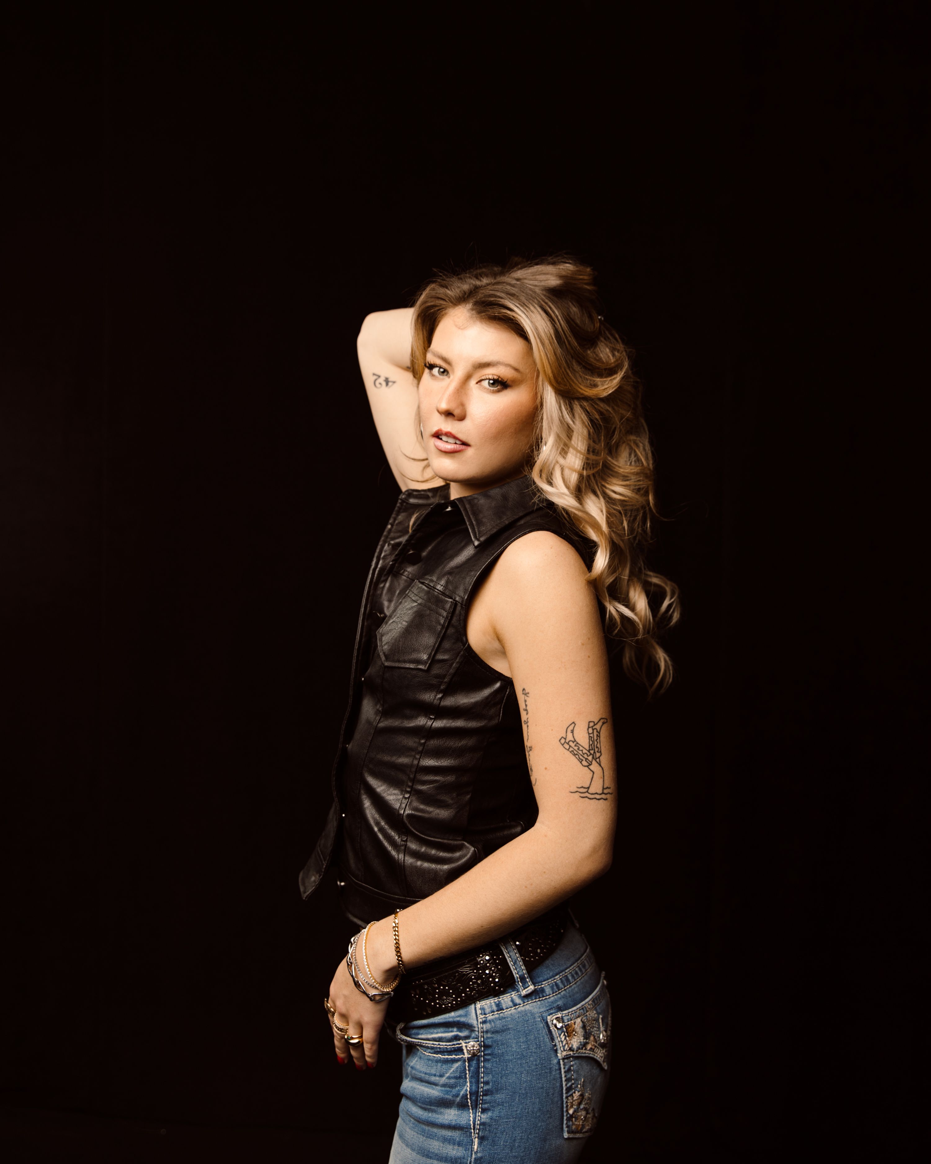 A person with wavy hair poses in a sleeveless leather vest and jeans, with tattoos visible on their arms.