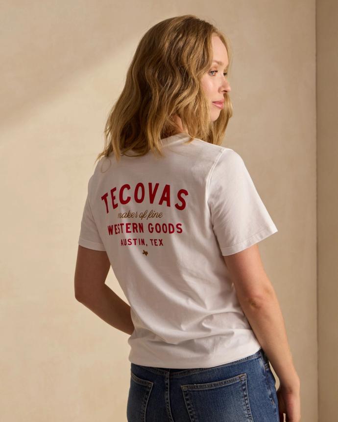 Woman wearing a white Tecovas t-shirt with red text that reads "Makers of Fine Western Goods, Austin, Tex," paired with blue jeans, standing sideways.