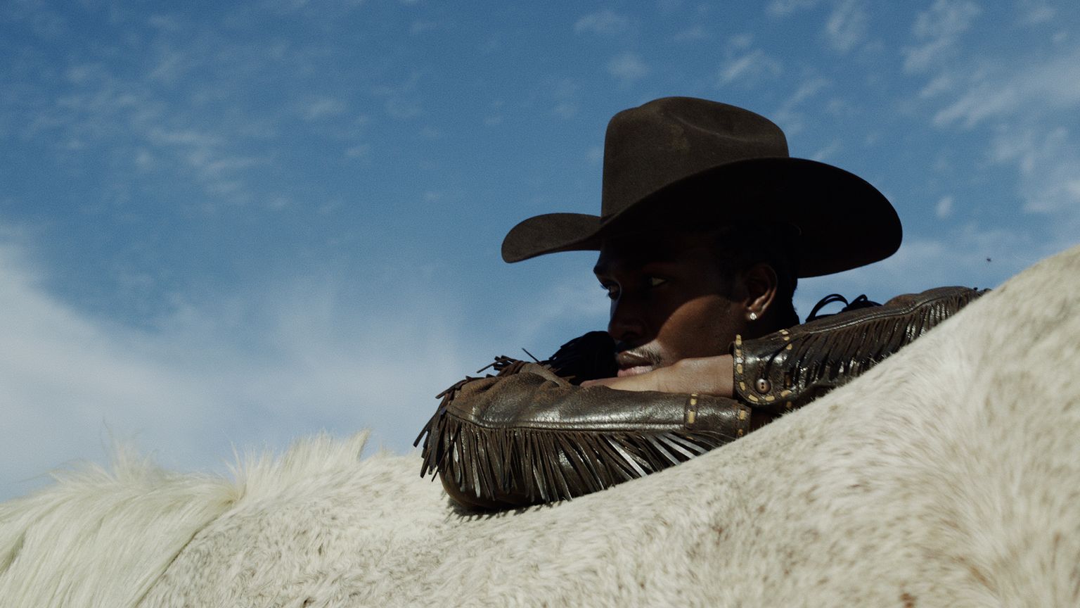 A person wearing a cowboy hat and fringed jacket leans on the back of a white horse under a blue sky.