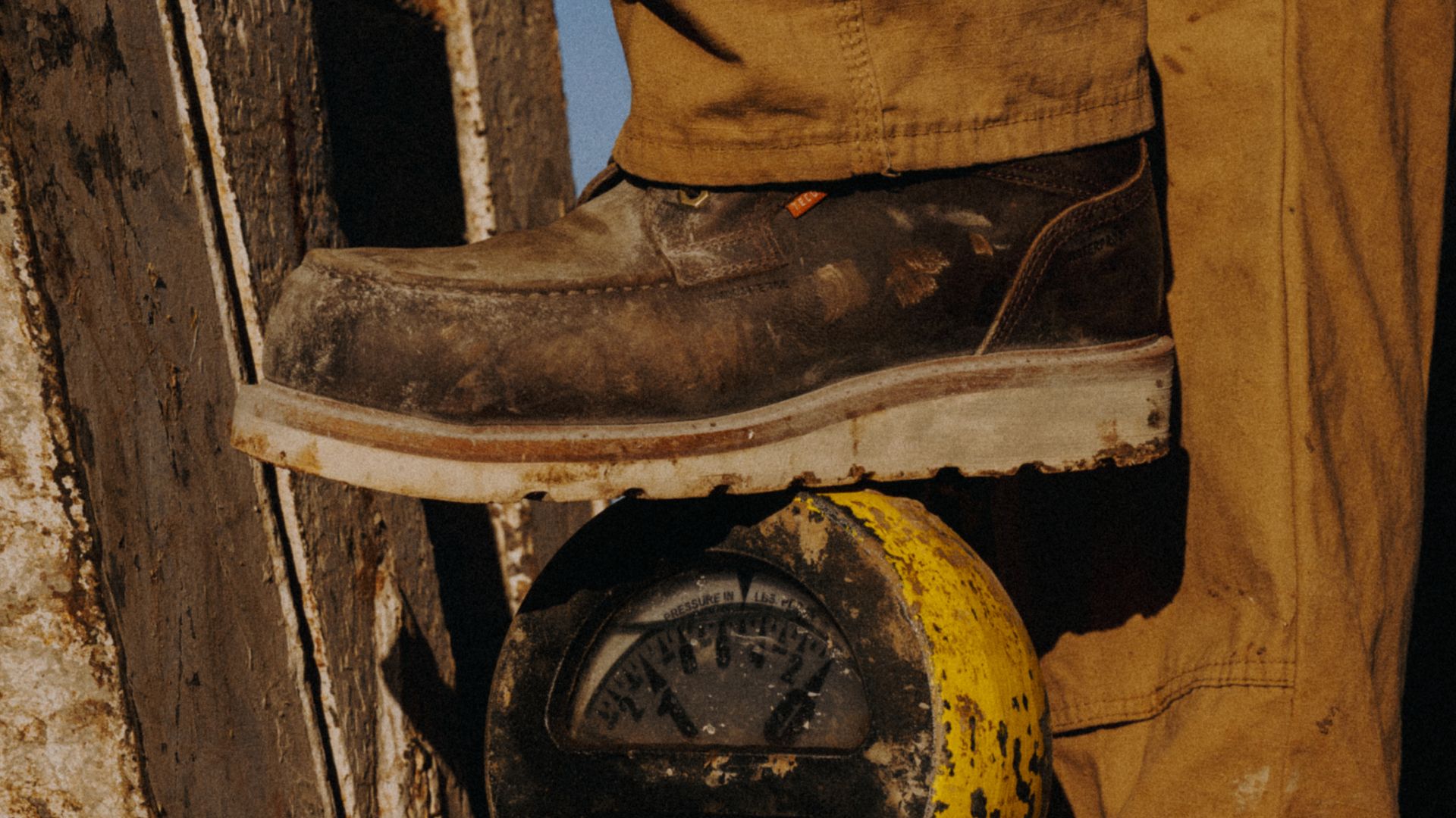 A person wearing a brown work boot and tan pants stands with one foot on a yellow industrial gauge.