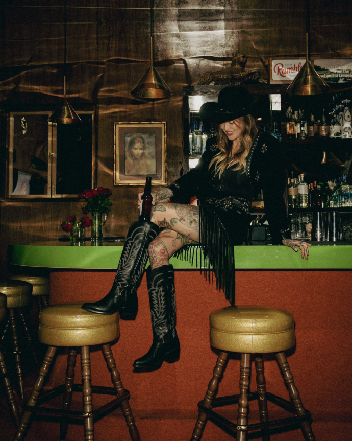 A woman in a cowboy hat and boots sits on a bar counter with a drink. The setting includes stools, a green bar top, and wooden wall decor.