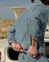 Person in a denim jacket and jeans stands near an open car trunk, adjusting their clothing with one hand, in an outdoor setting.