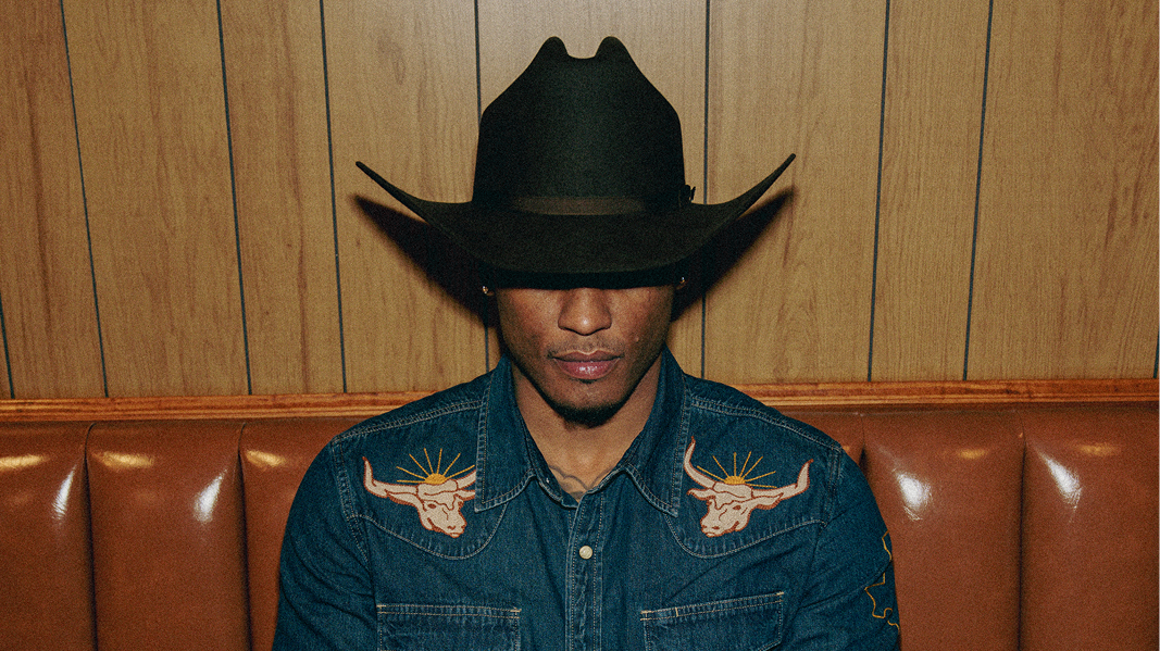 Person wearing a dark cowboy hat and denim shirt with bull designs sits in a brown booth with a wooden background.