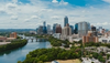 Panoramic view of a city skyline with a river running through it, featuring modern high-rise buildings, a bridge, and lush greenery under a partly cloudy sky.