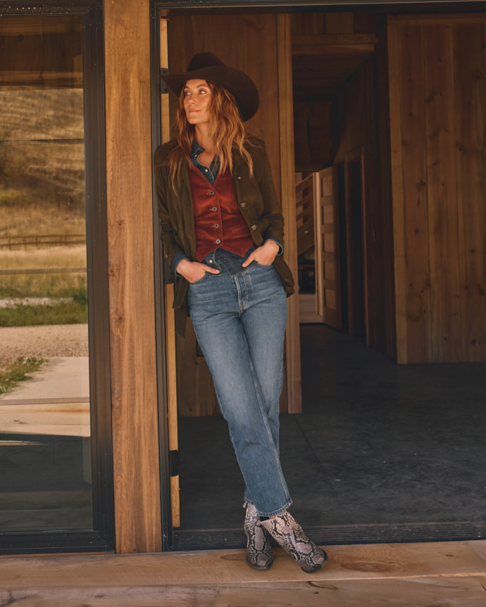 A woman in a cowboy hat, jeans, red vest, and snakeskin boots leans against a wooden doorway, looking to the side.