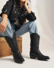 The Britt in Midnight Cowhide. A person in a black western shirt and blue jeans sits on a wooden box, wearing Midnight Cowhide cowboy boots with stitched detailing.