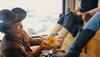 A person in a cowboy hat polishes white cowboy boots worn by another seated person, who is holding a drink can.
