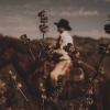 Dried plants in sharp focus in the foreground; a person wearing a cowboy hat rides a horse in the blurred background with a natural landscape.