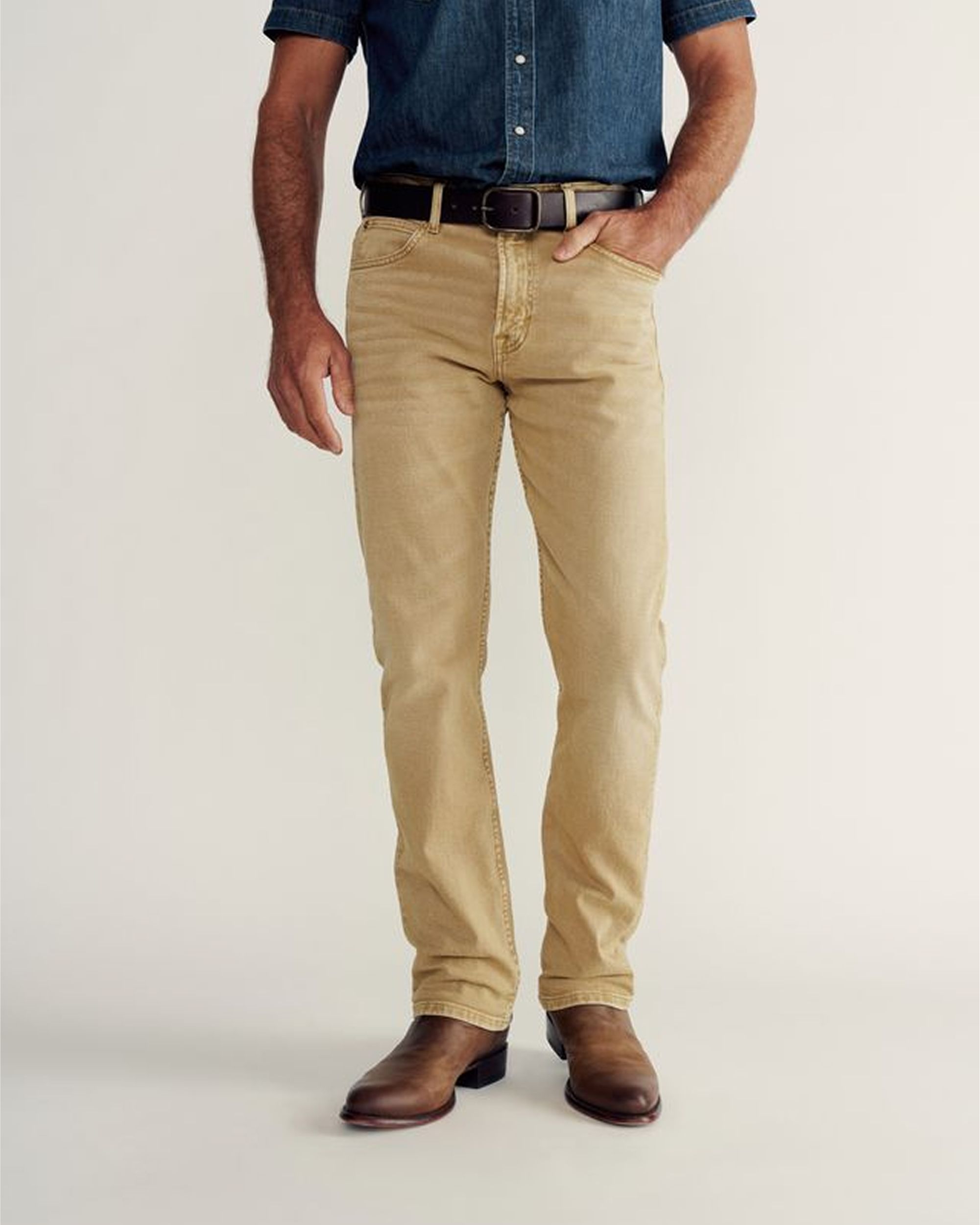 A person wearing a white t-shirt, olive green pants, and brown shoes stands with one hand in their pocket against a plain background.